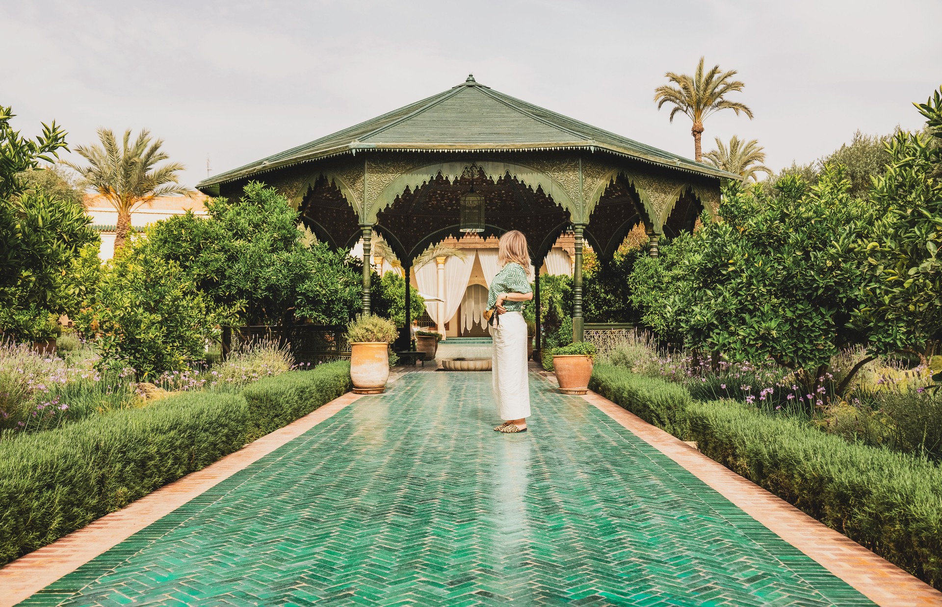 Young adult woman in stylish casual attire standing, looking away in a beautiful, serene lush green Moroccan Garden with traditional tiled path and gazebo, Le Jardin Secret, Marrakesh, Morocco